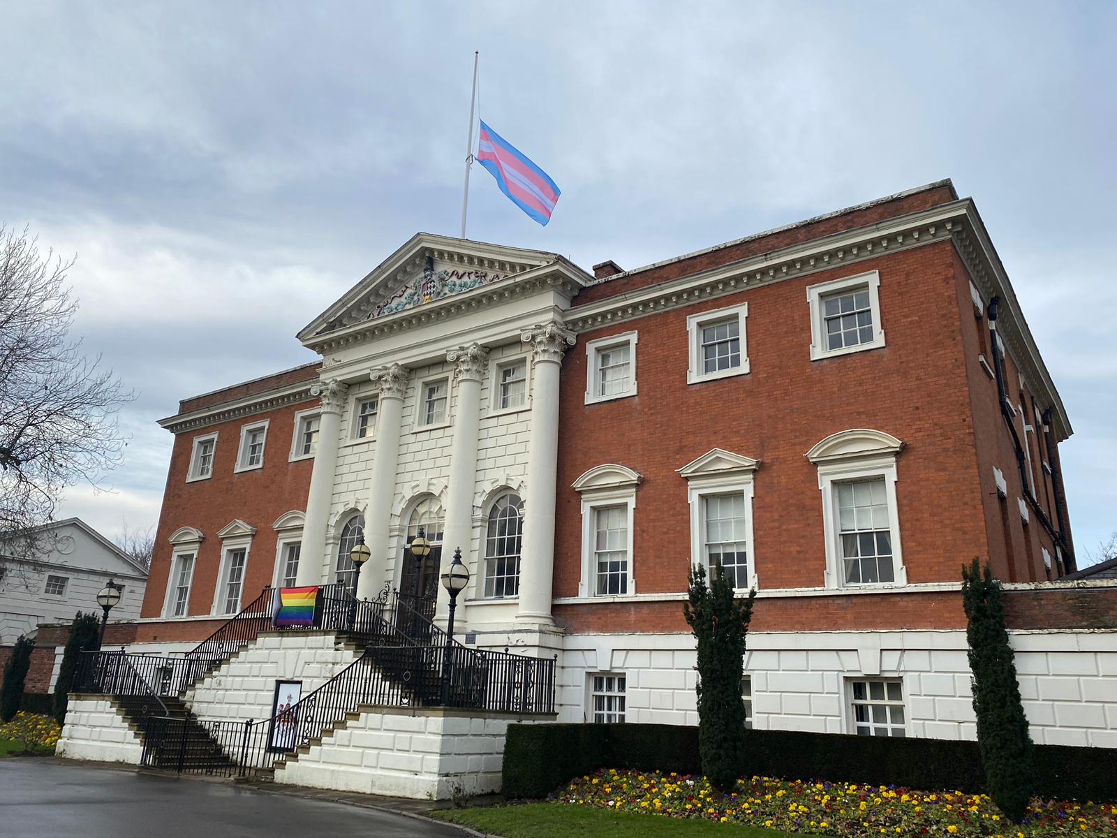 The Town Hall flag flying at half-mast with the trans flag on the flagpole and the progress pride flag affixed to the Town Hall railings