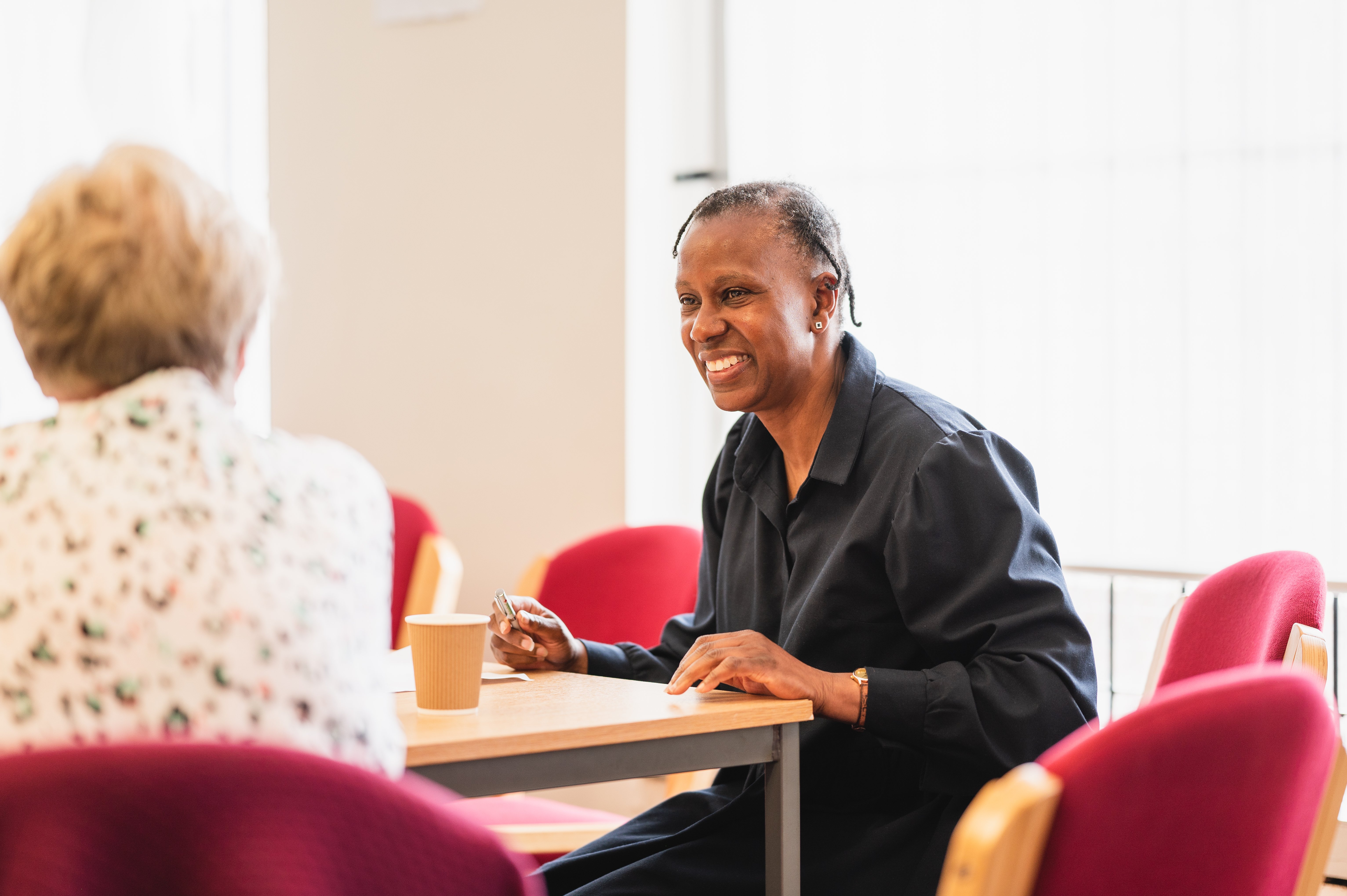 Image of two women sitting at a table and talking to each other, one woman has back to camera while other woman is facing camera