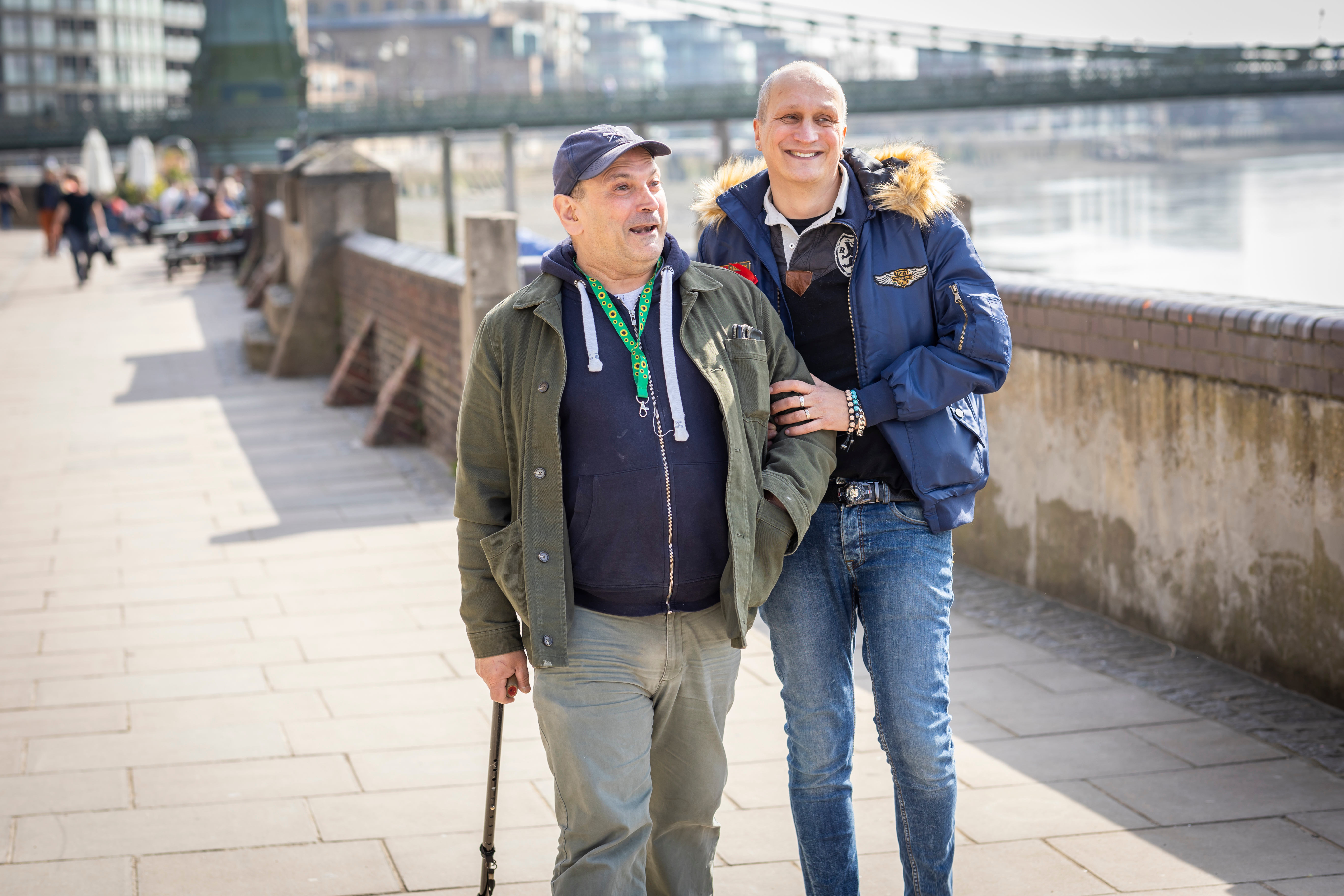 Two men walking along the seafront, one is walking with a cane, they are walking and linking arms. 