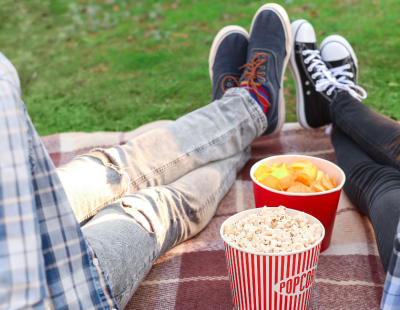 Photo of two people sat on a picnic blanket with a bucket of popcorn. 
